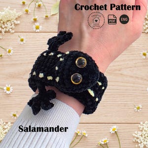 May include: A black crocheted salamander bracelet with small yellow spots and two large, round, dark eyes with gold-colored rims. The text reads Crochet Pattern, PDF, ENG, and Salamander. The item is wrapped around a wrist.