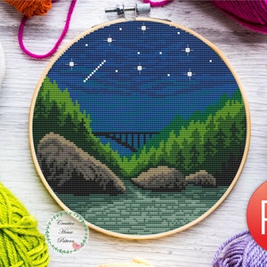 May include: Cross-stitch landscape scene in a wooden embroidery hoop. The design features a night sky with stars and a shooting star over a river and mountains. The PDF sticker is visible. Colorful yarn skeins surround the hoop.