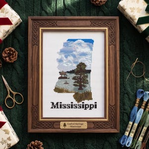 Può includere: Un ricamo a punto croce incorniciato del contorno dello stato del Mississippi, con un paesaggio con un albero e acqua sotto un cielo blu nuvoloso. La cornice è in legno scuro con angoli decorativi, e la parola "Mississippi" è ricamata sotto.