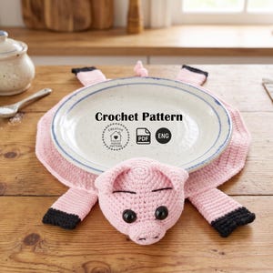 May include: A pink crocheted pig-shaped trivet with black accents, holding a white plate with blue trim. The trivet has a pig face and four legs. The text "Crochet Pattern" is visible on the plate.