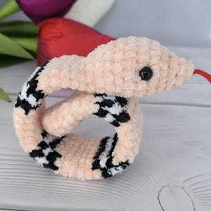 May include: A pink crochet pattern snake slap bracelet with black and white checkered accents. The snake has a red tongue and a black eye.