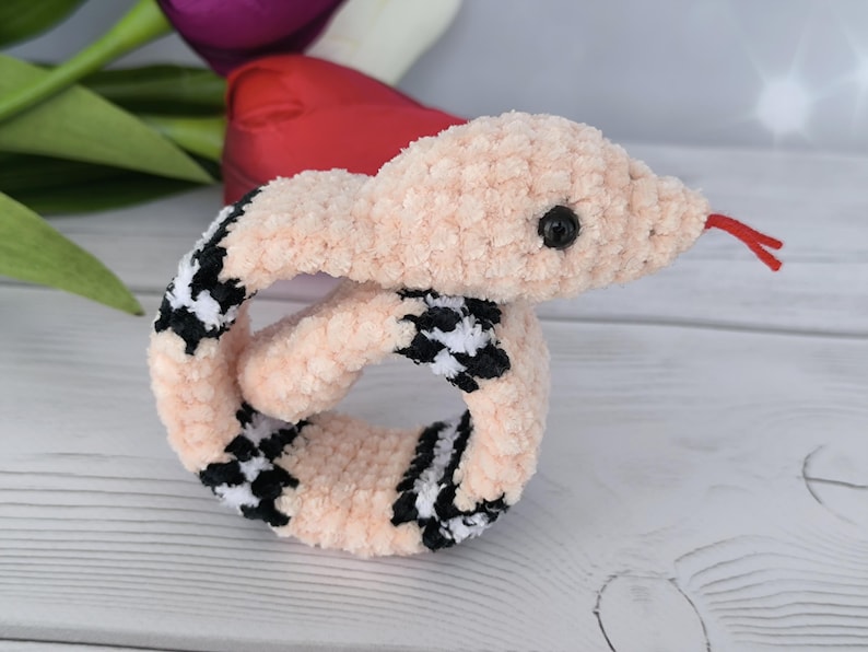 May include: A pink crochet pattern snake slap bracelet with black and white checkered accents. The snake has a red tongue and a black eye.