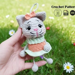 Peut inclure: Poup&eacute;e chat au crochet grise, orange et verte, avec un petit n&oelig;ud et des jambes pendantes. La poup&eacute;e a des yeux noirs et un nez rose. Le texte "Crochet Pattern" est visible en haut &agrave; droite.
