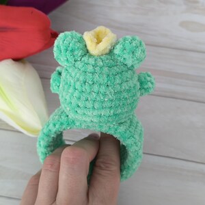 Peut inclure: Un bandeau en crochet vert clair avec un motif de grenouille en peluche sur le devant. La grenouille pr&eacute;sente une texture douce et duveteuse, de petites oreilles arrondies et une petite couronne jaune centr&eacute;e sur la t&ecirc;te en fil &eacute;pais.