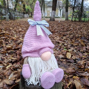 May include: A handmade crochet gnome doll with a pink hat and body, white beard, and a peach-colored nose. The gnome has a silver ribbon bow and is sitting on a wooden stump. The background is filled with fallen leaves.