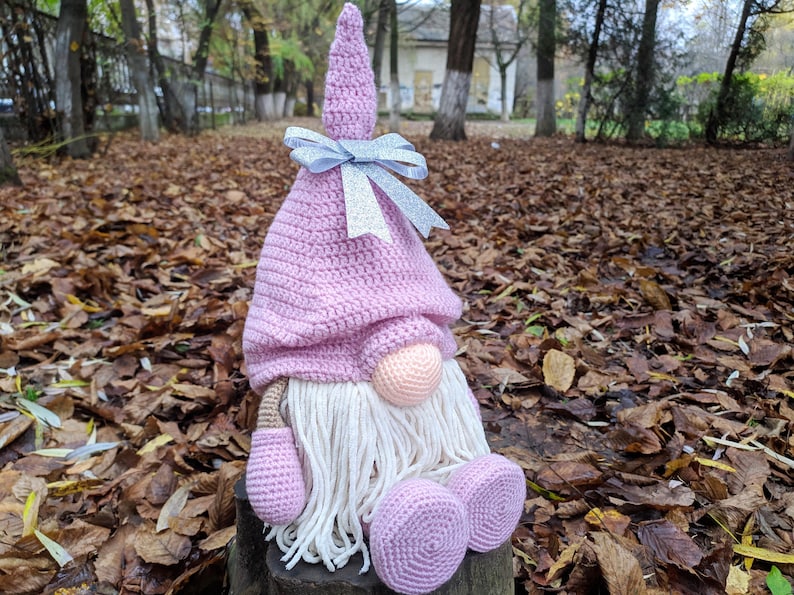 May include: A handmade crochet gnome doll with a pink hat and body, white beard, and a peach-colored nose. The gnome has a silver ribbon bow and is sitting on a wooden stump. The background is filled with fallen leaves.