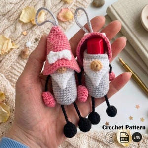 May include: Two handmade crochet gnome ornaments. One gnome has a pink hat with a white bow, a grey body, and a white beard. The other gnome has a pink hood, a grey body, and a white beard. Both have pink hands, black feet, and grey hanging loops.