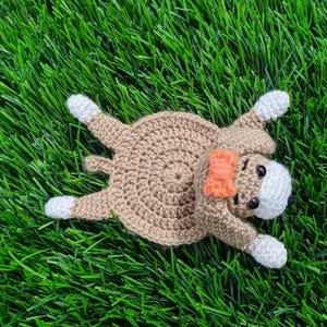 May include: A handmade crochet dog coaster in a lying-down pose. The coaster is primarily tan with white paws and a white face. It has a small orange bow and black eyes. The coaster is lying on a bed of green grass.
