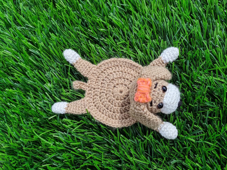 May include: A handmade crochet dog coaster in a lying-down pose. The coaster is primarily tan with white paws and a white face. It has a small orange bow and black eyes. The coaster is lying on a bed of green grass.