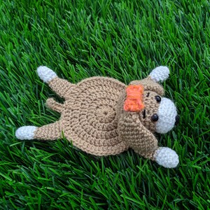 May include: A handmade crochet dog coaster laying on green grass. The dog is brown with white paws and a white face. It has black eyes and an orange bow. The dog is a whimsical, handcrafted item.