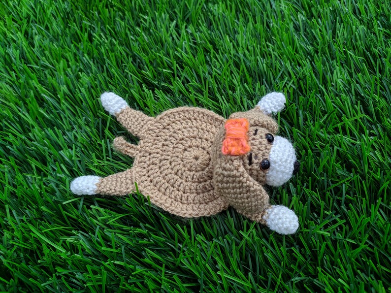 May include: A handmade crochet dog coaster laying on green grass. The dog is brown with white paws and a white face. It has black eyes and an orange bow. The dog is a whimsical, handcrafted item.