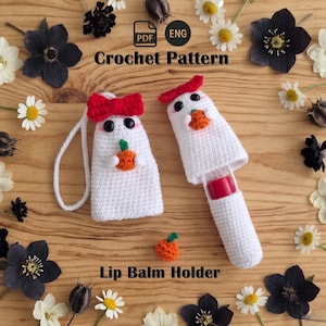 May include: Two white crocheted ghost-shaped lip balm holders with black eyes and pink cheeks. One ghost has a red bow, the other a red hat, and both hold a small orange pumpkin. Text reads: PDF ENG Crochet Pattern Lip Balm Holder.