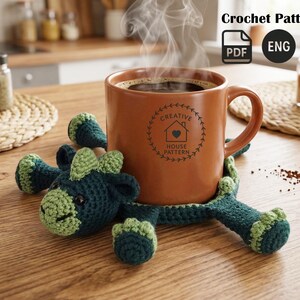 Puede incluir: Una taza de cerámica marrón llena de café descansa sobre un posavasos de ganchillo verde y azul hecho a mano con forma de dragón. La taza tiene un logotipo que dice "Creative House Pattern". Sale vapor del café. Las palabras "Crochet Pattern" y "ENG" están en la esquina superior derecha.