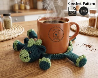 May include: A brown ceramic mug filled with coffee sits on a handmade, green and blue crochet coaster shaped like a dragon. The mug has a logo that says "Creative House Pattern". Steam rises from the coffee. The image also includes the text "Crochet Pattern" and "ENG".