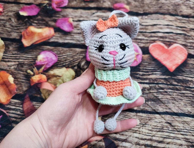 Peut inclure: Lapin crochet&eacute; gris avec un n&oelig;ud orange, un nez rose et des yeux noirs. Le lapin porte une tenue verte et orange avec une jupe vert clair. Il est fait &agrave; la main et a des jambes pendantes avec des pompons gris.