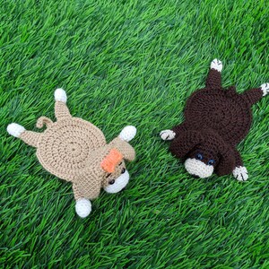 "A top-down view of two crocheted coasters shaped like dogs laying on a green grass surface. On the left is a tan crocheted dog coaster wearing an orange bow. On the right is a dark brown crocheted dog coaster. Both coasters feature small crocheted bodies, floppy ears, white paws, small safety eyes, and little white muzzles. These handmade items are based on a crochet pattern for dog coasters."