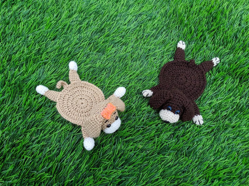 "A top-down view of two crocheted coasters shaped like dogs laying on a green grass surface. On the left is a tan crocheted dog coaster wearing an orange bow. On the right is a dark brown crocheted dog coaster. Both coasters feature small crocheted bodies, floppy ears, white paws, small safety eyes, and little white muzzles. These handmade items are based on a crochet pattern for dog coasters."