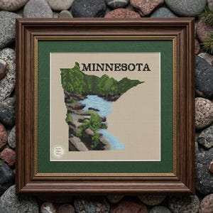 May include: Framed cross-stitch art featuring the outline of Minnesota, with a waterfall and lush greenery. The word "MINNESOTA" is stitched above the state's shape. The artwork is in a brown wooden frame with a green mat.