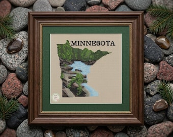 May include: Framed cross-stitch art featuring the outline of Minnesota, with a waterfall and lush greenery. The word "MINNESOTA" is stitched above the state's shape. The artwork is in a brown wooden frame with a green mat.