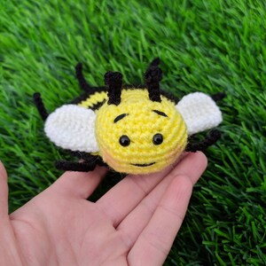 May include: A handmade crochet bee toy with a yellow body, black and white stripes, and white wings. The bee has black eyes and antennae. The toy is lying on a bed of green artificial grass.