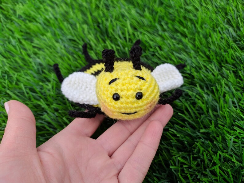 May include: A handmade crochet bee toy with a yellow body, black and white stripes, and white wings. The bee has black eyes and antennae. The toy is lying on a bed of green artificial grass.