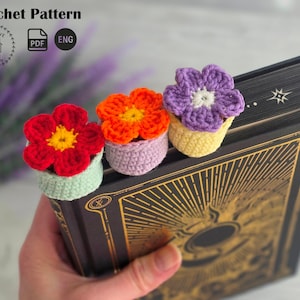 May include: Three colorful crochet flower bookmarks rest on a dark book. The flowers are red, orange, and purple, each with a yellow center. The bookmarks are in small, pot-like holders. The image includes the text "Crochet Pattern".