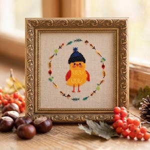 May include: A framed cross-stitch featuring a yellow bird wearing a blue hat, surrounded by a wreath of leaves and acorns. The frame is gold-colored with ornate detailing. The image is set against a natural background with fall foliage.