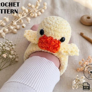 May include: A crocheted yellow duck with an orange beak and black eyes. The duck is handmade and has a fluffy texture. The image also includes the text "CROCHET PATTERN".