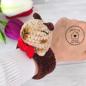 May include: Sensory Capybara Fidget Toy Crochet Pattern with a red bow tie. The Capybara has closed eyes and is attached to a dark brown wrist cuff. The wristband is shown on a person's wrist.