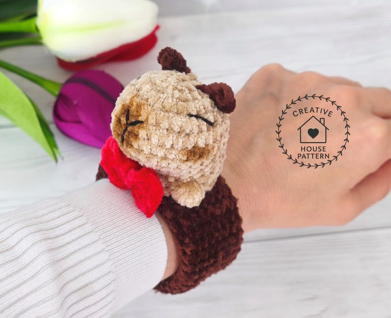 May include: Sensory Capybara Fidget Toy Crochet Pattern with a red bow tie. The Capybara has closed eyes and is attached to a dark brown wrist cuff. The wristband is shown on a person's wrist.