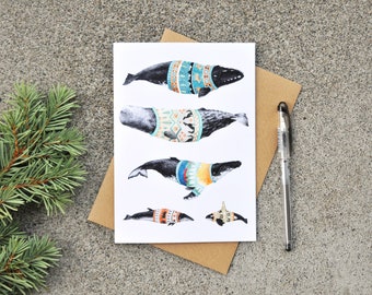 May include: A white card with a colorful illustration of five whales wearing sweaters. The whales are different sizes and have different patterns on their sweaters. The card is on a brown surface.