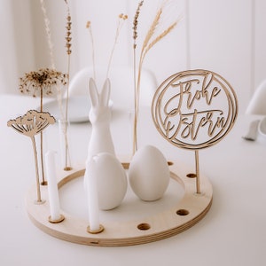 Könnte beinhalten: Ein rundes Holz-Oster-Mittelstück mit 12 Löchern. Die Basis hat einen weißen Keramik-Hasen, zwei weiße Keramik-Eier und ein Holzschild mit der Aufschrift "Frohe Ostern".