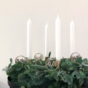 Könnte beinhalten: Ein weißer Adventskranz mit vier weißen Kerzen und Holzschmuck mit den Zahlen "eins", "zwei", "drei" und "vier" darauf.