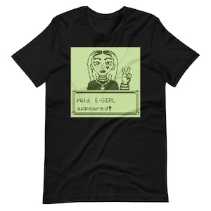 May include: Black t-shirt with a green pixelated image of a woman with pink hearts on her cheeks. The image is in a green box with the text "Wild E-GIRL appeared!"