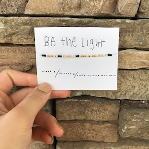 May include: A white card with the text "Be the Light" written in black marker. A beaded bracelet with black and gold beads is strung across the card.