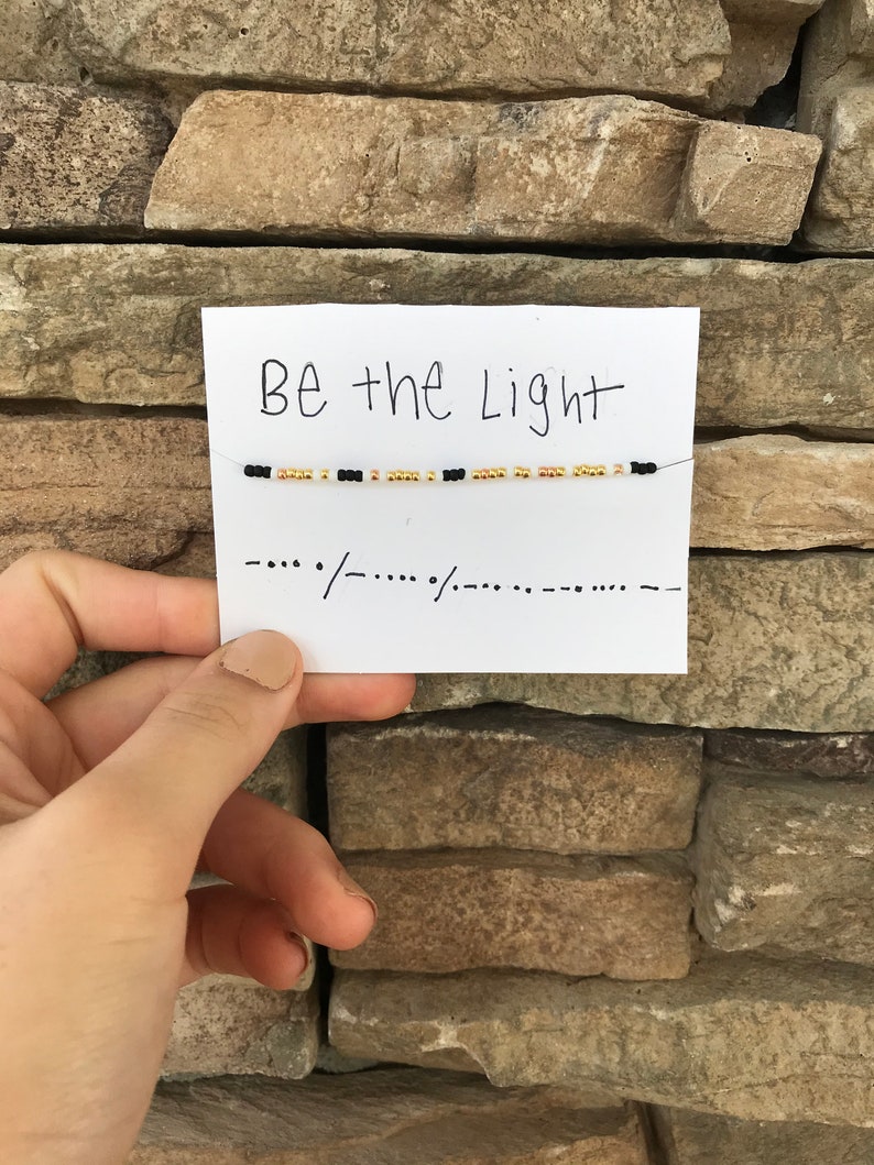 May include: A white card with the text "Be the Light" written in black marker. A beaded bracelet with black and gold beads is strung across the card.