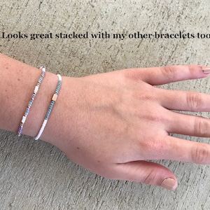 May include: Two beaded bracelets stacked on a wrist. The top bracelet is white with small blue beads. The bottom bracelet is white with small blue, purple, and gold beads.