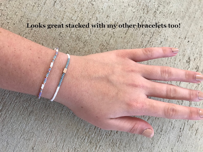 May include: Two beaded bracelets stacked on a wrist. The top bracelet is white with small blue beads. The bottom bracelet is white with small blue, purple, and gold beads.