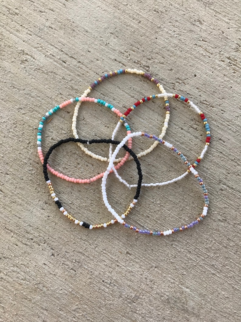 May include: Six colorful beaded bracelets in various colors including black, white, pink, blue, and gold. The bracelets are made of small beads and are stacked on top of each other.