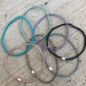 May include: A set of seven beaded bracelets in various colors, including turquoise, light blue, lavender, gray, and black. Each bracelet has a small gold accent.