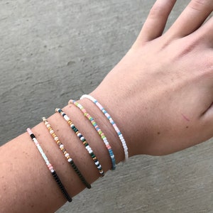 May include: A close-up of a person's wrist wearing six beaded bracelets. The bracelets are made of small, colorful beads in various patterns. The bracelets are stacked on top of each other, creating a layered look.