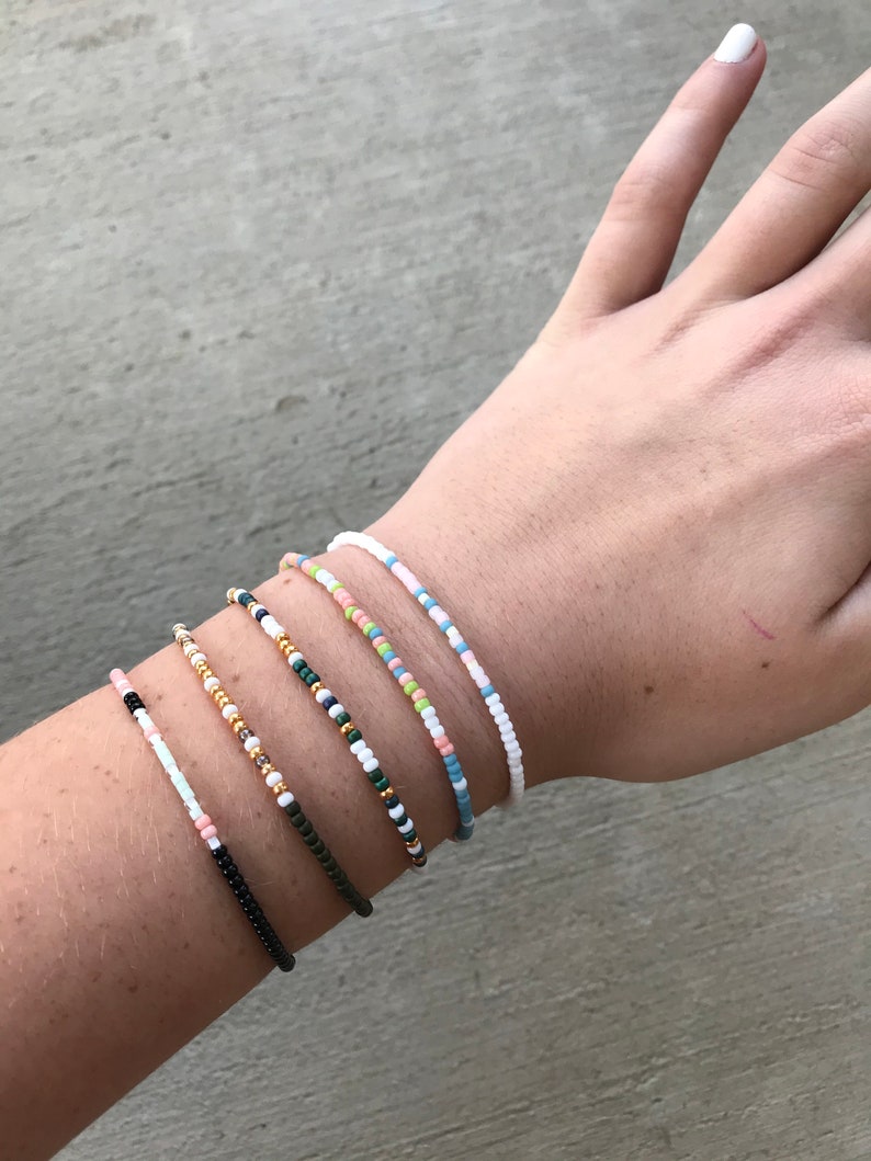 May include: A close-up of a person's wrist wearing six beaded bracelets. The bracelets are made of small, colorful beads in various patterns. The bracelets are stacked on top of each other, creating a layered look.