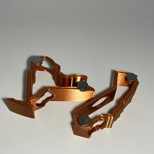 Puede incluir: Dos clips magn&eacute;ticos impresos en 3D, de color cobre, con un dise&ntilde;o decorativo. Cada clip tiene un peque&ntilde;o im&aacute;n redondo plateado en la parte inferior.