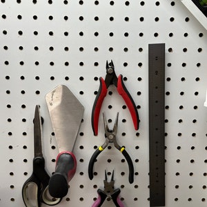 May include: A white pegboard with a variety of tools hanging on it, including a pair of red and black wire cutters, a pair of yellow and black pliers, a pair of purple pliers, a pair of black scissors, a silver putty knife, and a black ruler marked with numbers 1 through 11.