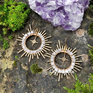Pu&ograve; includere: Un paio di orecchini dorati a forma di raggio di sole con una stella al centro. Gli orecchini hanno un anello di perline bianche. Lo sfondo &egrave; una pietra naturale con muschio verde e un cristallo viola.