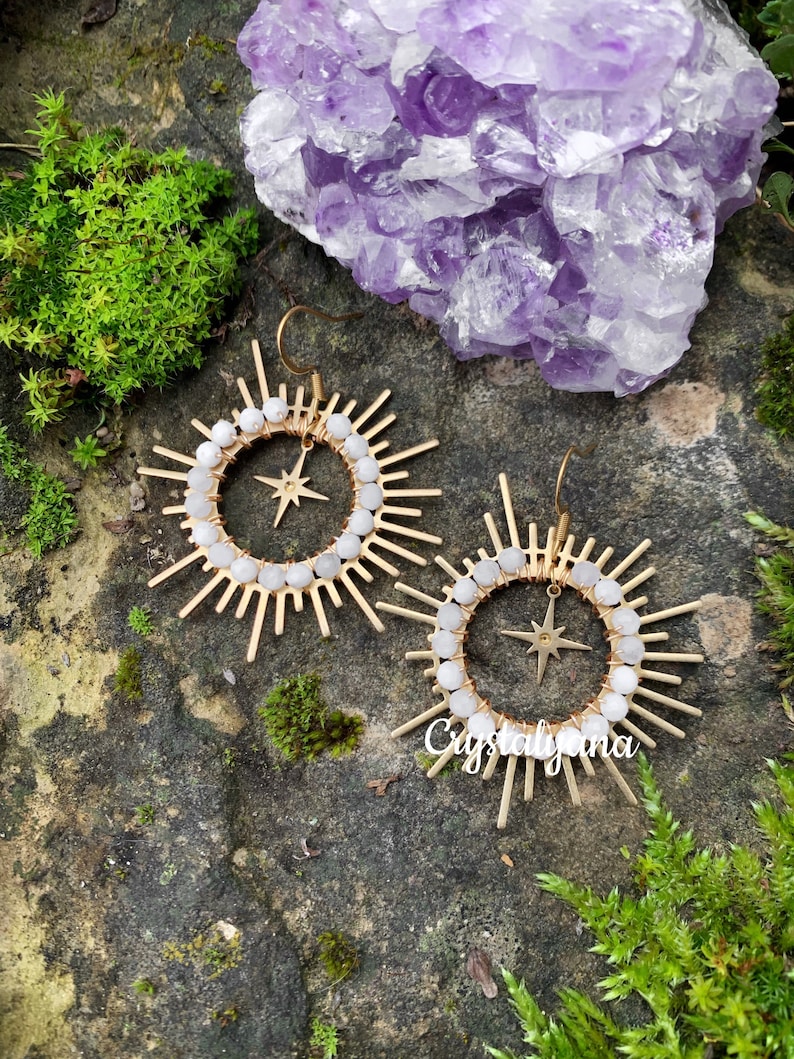 Pu&ograve; includere: Un paio di orecchini dorati a forma di raggio di sole con una stella al centro. Gli orecchini hanno un anello di perline bianche. Lo sfondo &egrave; una pietra naturale con muschio verde e un cristallo viola.