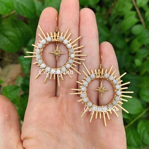 Pu&ograve; includere: Un paio di orecchini color oro con un design a raggiera. Ogni orecchino presenta una stella centrale, circondata da un cerchio di perline bianche e barre dorate radianti. Gli orecchini sono esposti su uno sfondo verde.