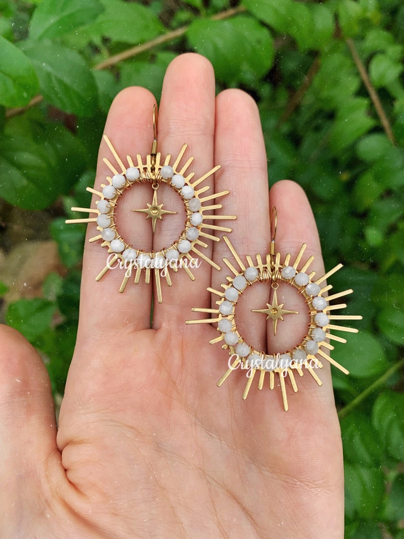 Pu&ograve; includere: Un paio di orecchini color oro con un design a raggiera. Ogni orecchino presenta una stella centrale, circondata da un cerchio di perline bianche e barre dorate radianti. Gli orecchini sono esposti su uno sfondo verde.