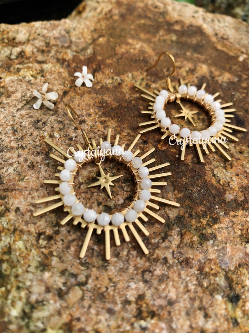 Pu&ograve; includere: Un paio di orecchini dorati a forma di sole con perline bianche che circondano un centro a forma di stella. Gli orecchini hanno una chiusura a gancio e sono esposti su una superficie marrone strutturata. Il testo "Cristalyana" &egrave; visibile.