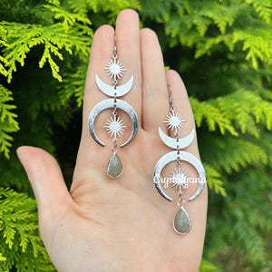 May include: Silver-toned celestial earrings featuring crescent moons, starbursts, and teardrop-shaped gemstones. The earrings are displayed against a backdrop of green foliage, showcasing their intricate design and elegant style.